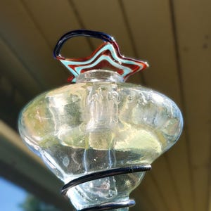 May include: A clear glass decorative piece with a star-shaped top in red, blue, and turquoise. The piece has a bulbous body and a black wire base. The top has a curved handle in blue. The background is a blurred view of a light-colored surface.