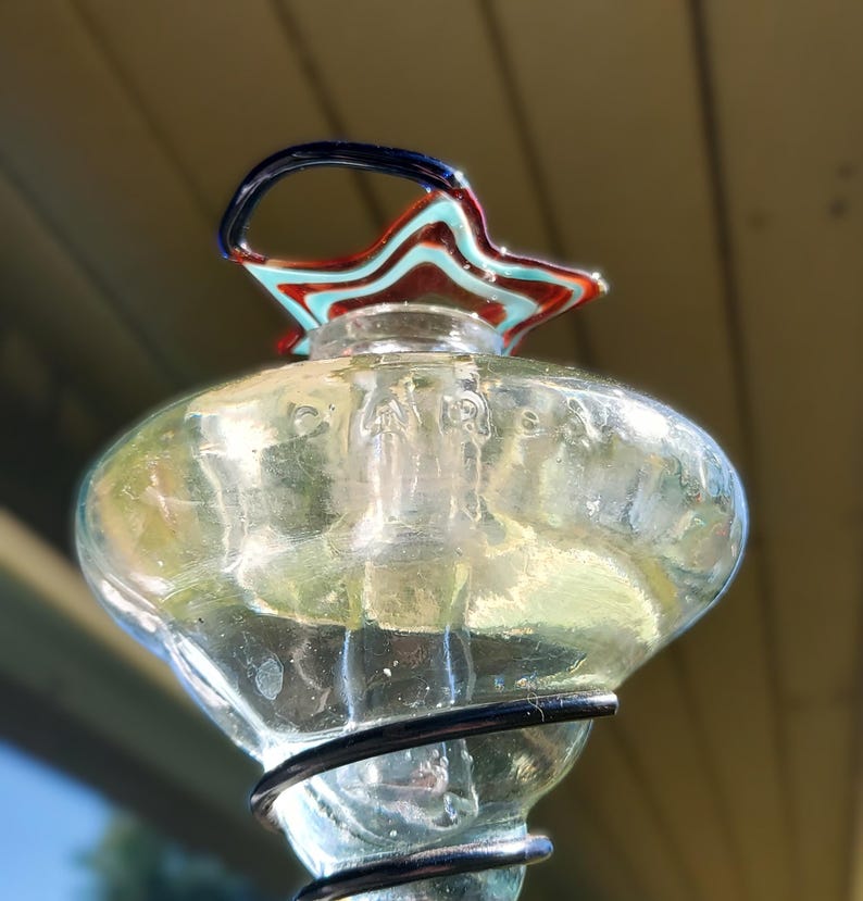 May include: A clear glass decorative piece with a star-shaped top in red, blue, and turquoise. The piece has a bulbous body and a black wire base. The top has a curved handle in blue. The background is a blurred view of a light-colored surface.