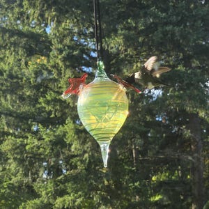 May include: A glass hummingbird feeder with a teardrop shape, filled with liquid. The feeder has red star-shaped perches and a green top. A hummingbird is feeding from the feeder, with its wings spread. The background is a blurred view of green trees.
