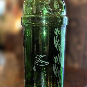 May include: A green glass container with a decorative design. The container features a hummingbird and tulip design etched in white. The container is cylindrical with a rounded top and a small, circular opening.