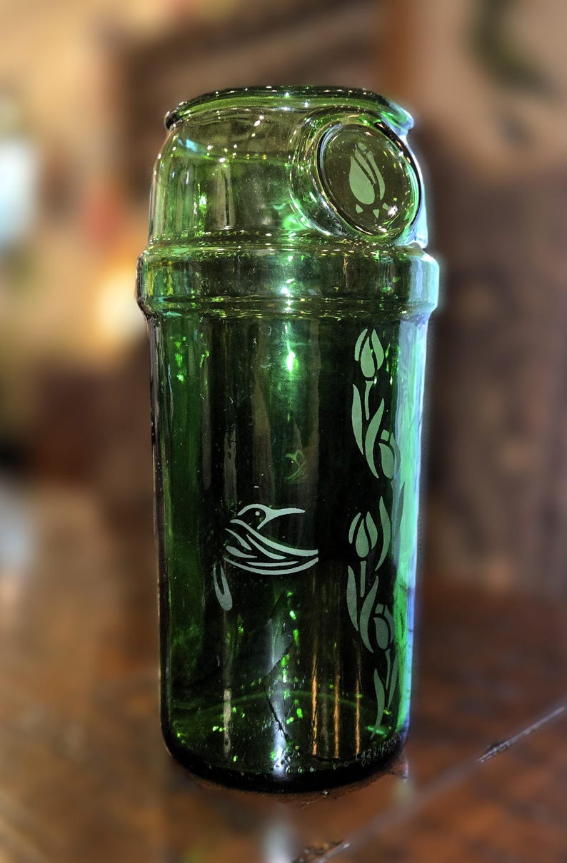 May include: A green glass container with a decorative design. The container features a hummingbird and tulip design etched in white. The container is cylindrical with a rounded top and a small, circular opening.
