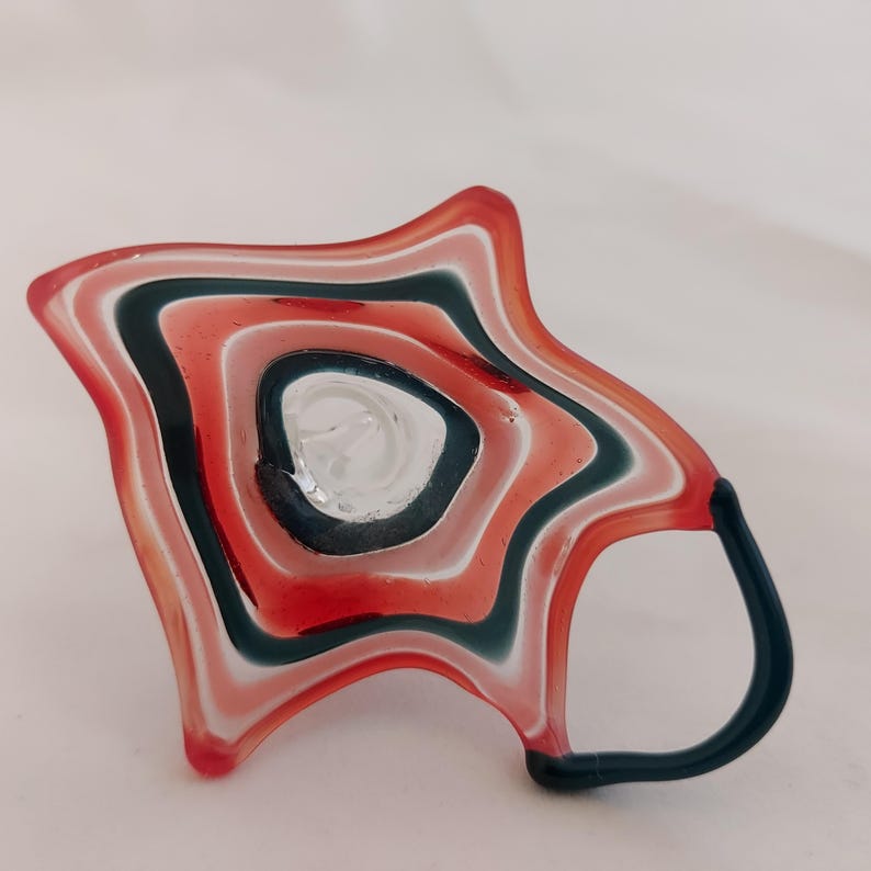 May include: A unique, handmade glass mug with a star-like shape. The mug features a layered design of red, white, and dark teal stripes. The handle is dark teal. The interior of the mug is clear glass.