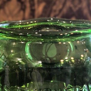 May include: Close-up of a green glass bottle. The bottle has a rounded top with a slightly wavy rim. The glass reflects light, creating highlights and shadows. The bottle's surface appears smooth and clear.