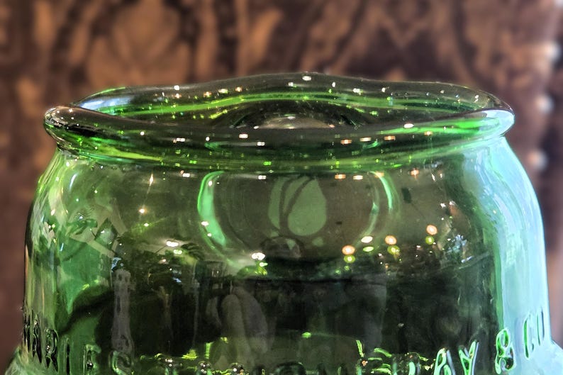 May include: Close-up of a green glass bottle. The bottle has a rounded top with a slightly wavy rim. The glass reflects light, creating highlights and shadows. The bottle's surface appears smooth and clear.