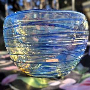 May include: A clear glass bowl with a rounded shape, featuring horizontal blue and yellow swirls. The bowl has a glossy finish and is set against a blurred background.