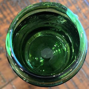 May include: An overhead view of a dark green glass container. The glass has a glossy finish and a slightly textured surface. The interior of the container is visible, with a circular base and a wider opening. The container is likely a drinking glass or vase.