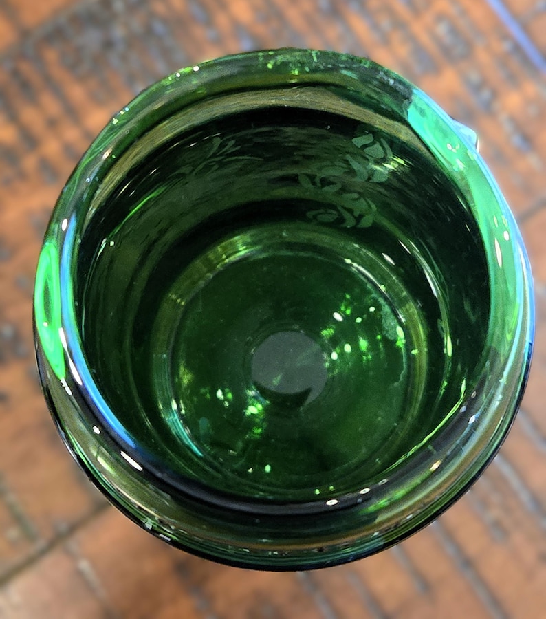 May include: An overhead view of a dark green glass container. The glass has a glossy finish and a slightly textured surface. The interior of the container is visible, with a circular base and a wider opening. The container is likely a drinking glass or vase.