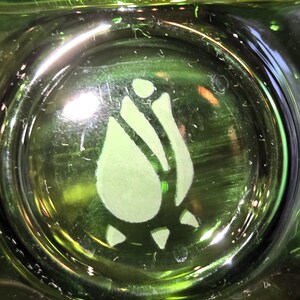 May include: Close-up of a green glass object with a light green rose design etched in the center. The glass has a glossy finish and is illuminated, creating highlights and reflections. The rose design is a stylized floral element.