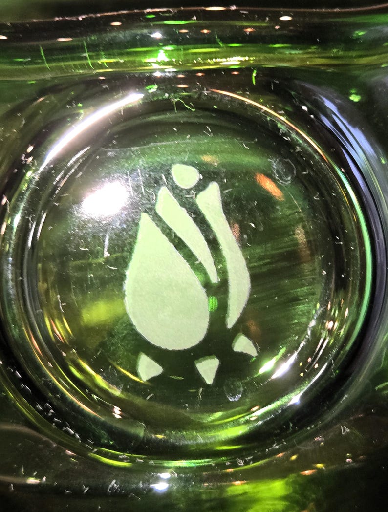 May include: Close-up of a green glass object with a light green rose design etched in the center. The glass has a glossy finish and is illuminated, creating highlights and reflections. The rose design is a stylized floral element.