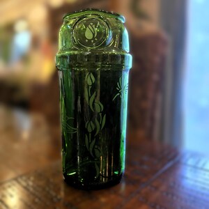 May include: A green glass cocktail shaker with etched floral and insect designs. The shaker has a cylindrical body and a rounded top with a circular emblem. The glass is a deep, rich green color, and the etched designs add a decorative touch.