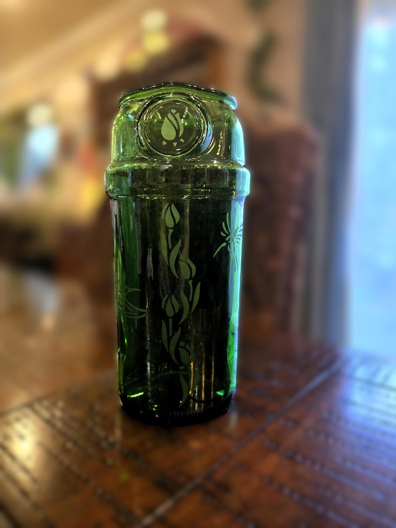 May include: A green glass cocktail shaker with etched floral and insect designs. The shaker has a cylindrical body and a rounded top with a circular emblem. The glass is a deep, rich green color, and the etched designs add a decorative touch.