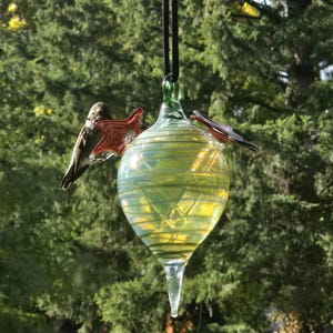 May include: A hanging glass hummingbird feeder with a teardrop shape, featuring a green and yellow iridescent finish. Two red glass hummingbird perches are attached. The feeder hangs from a black cord against a backdrop of green trees.