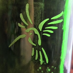 May include: A green glass bottle with a neon green hummingbird design. The hummingbird is depicted in a simple, stylized form, with its wings spread. The bottle's surface is reflective, and the design is illuminated.
