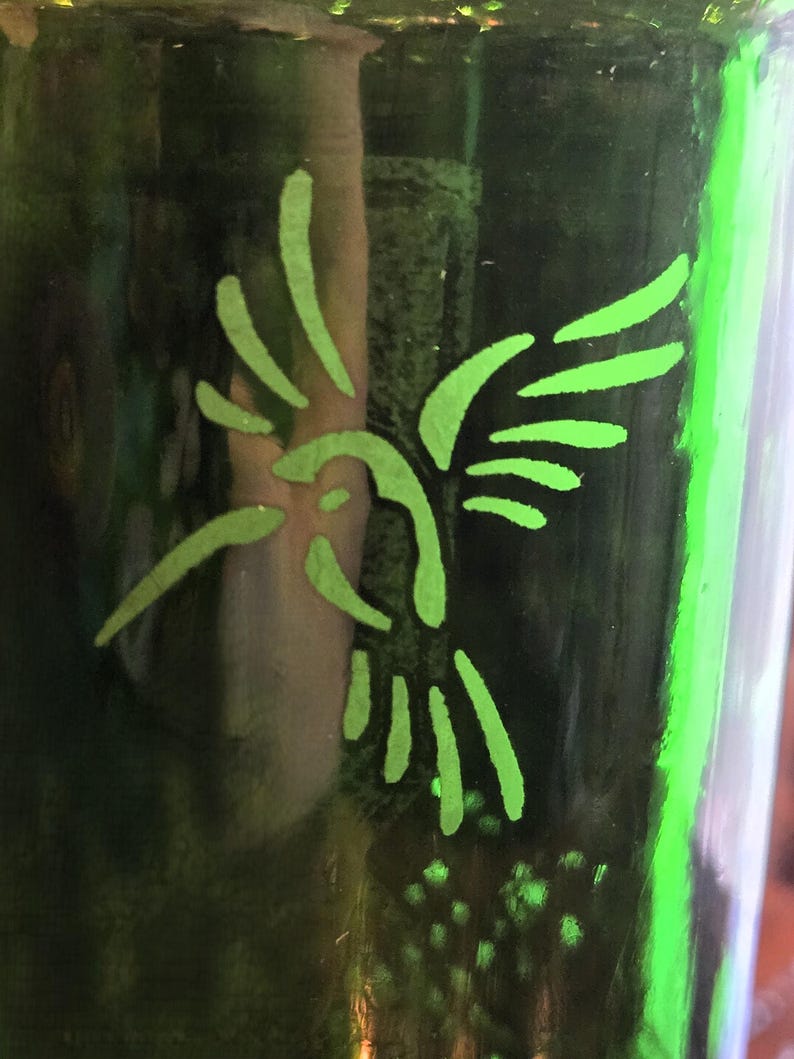 May include: A green glass bottle with a neon green hummingbird design. The hummingbird is depicted in a simple, stylized form, with its wings spread. The bottle's surface is reflective, and the design is illuminated.