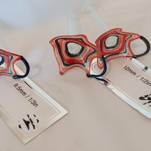 May include: Two abstract glass ornaments with red, black, and white patterns. One is labeled 9.5mm / 1/3in, the other 12mm / 1/2in. Each has a dark blue handle and a clear stem.