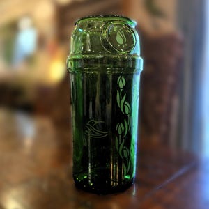 May include: A green glass beverage container with a decorative design. The container features etched images of a bird and floral elements. The top has a circular design with a floral motif. The container is made of green glass.