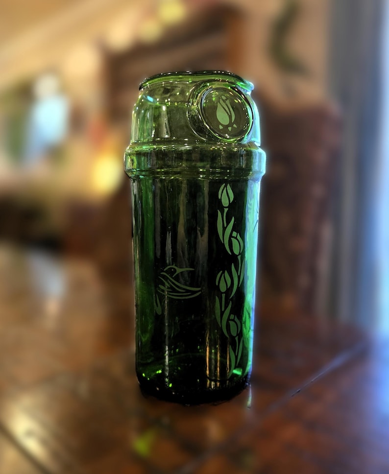 May include: A green glass beverage container with a decorative design. The container features etched images of a bird and floral elements. The top has a circular design with a floral motif. The container is made of green glass.