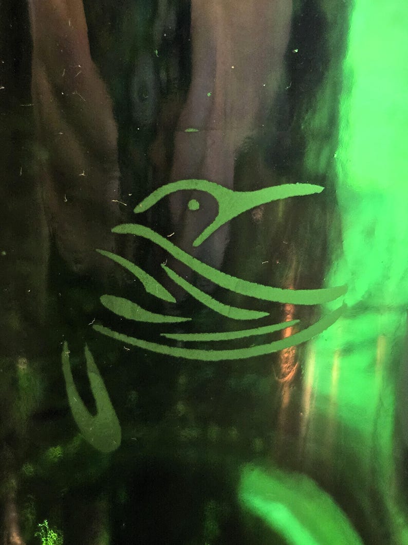 May include: A close-up of a green glass surface with a stylized hummingbird design etched into it. The hummingbird is depicted in a simple, elegant style, with flowing lines representing its wings and body. The background is a dark green.