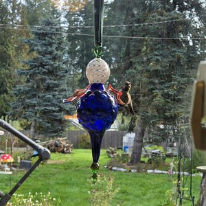 May include: A handcrafted glass bird feeder with a deep blue body, a white and clear patterned globe, and red and orange accents. A small hummingbird perches on the feeder. The feeder hangs from a green ribbon against a backdrop of trees.