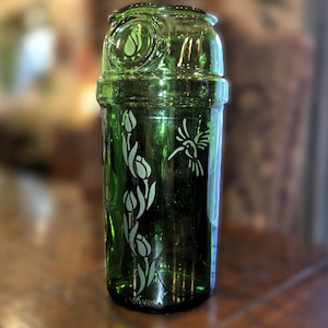 May include: A green glass cocktail shaker with etched floral and hummingbird designs. The shaker has a cylindrical body with a rounded top and a decorative emblem. The etched designs are white, contrasting against the green glass.
