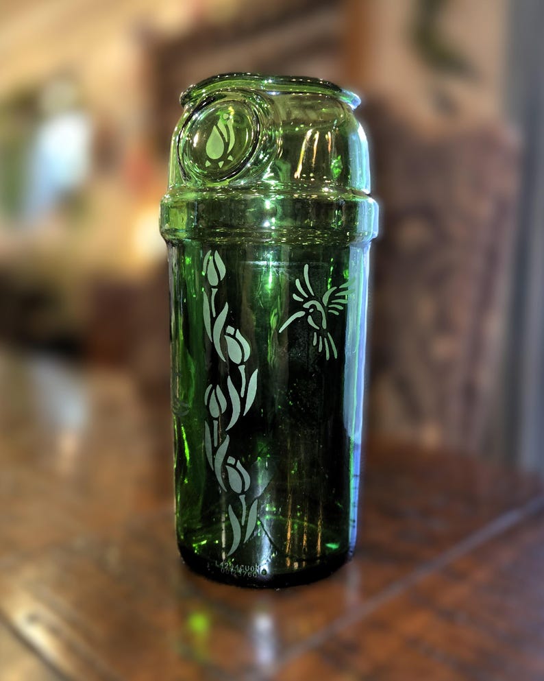 May include: A green glass cocktail shaker with etched floral and hummingbird designs. The shaker has a cylindrical body with a rounded top and a decorative emblem. The etched designs are white, contrasting against the green glass.