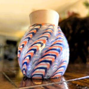 May include: A decorative glass vase with a white top and a bulbous body. The vase features a swirling pattern of blue, orange, and white stripes. The vase is sitting on a wooden surface.