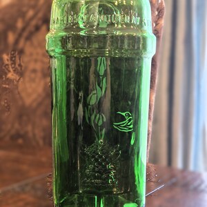 May include: A vintage green glass bottle with embossed floral and bird designs. The bottle has a cylindrical shape with a wide mouth and a slightly raised band around the middle. The text "HAWKES & ANTOINE" is embossed near the top.