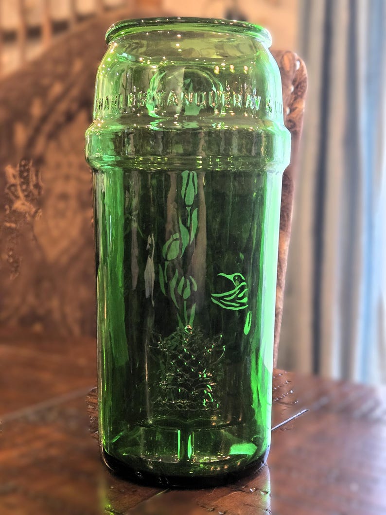 May include: A vintage green glass bottle with embossed floral and bird designs. The bottle has a cylindrical shape with a wide mouth and a slightly raised band around the middle. The text "HAWKES & ANTOINE" is embossed near the top.