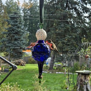 May include: A handcrafted glass hummingbird feeder. The feeder features a deep blue, teardrop-shaped body with red and orange accents, topped with a clear, swirled glass globe. A hummingbird is perched on the feeder, feeding. The feeder hangs from a green ribbon.