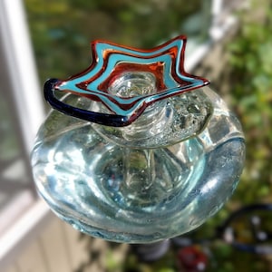 May include: A clear glass bottle stopper with a star-shaped top. The star features a teal and red swirl design. The stopper has a dark blue handle and is set against a blurred green background.