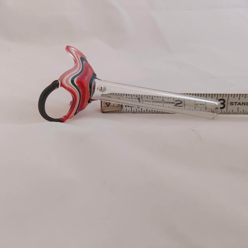 May include: A clear glass tool with a decorative handle featuring red, white, and blue swirls and a black ring. The tool is approximately 3 inches long. The tool is placed on a white surface.