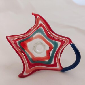 May include: A star-shaped glass object with a handle. The object features red, white, teal, and pink swirls. The handle is a solid dark blue. The object is likely a decorative piece or a small container.