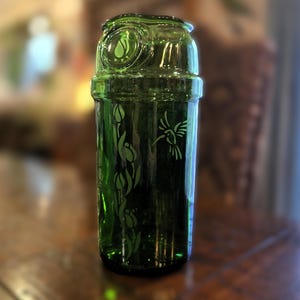 May include: A green glass bottle with a decorative design. The bottle features etched images of flowers and a hummingbird. The top of the bottle has a circular design with a leaf. The bottle is sitting on a wooden surface.