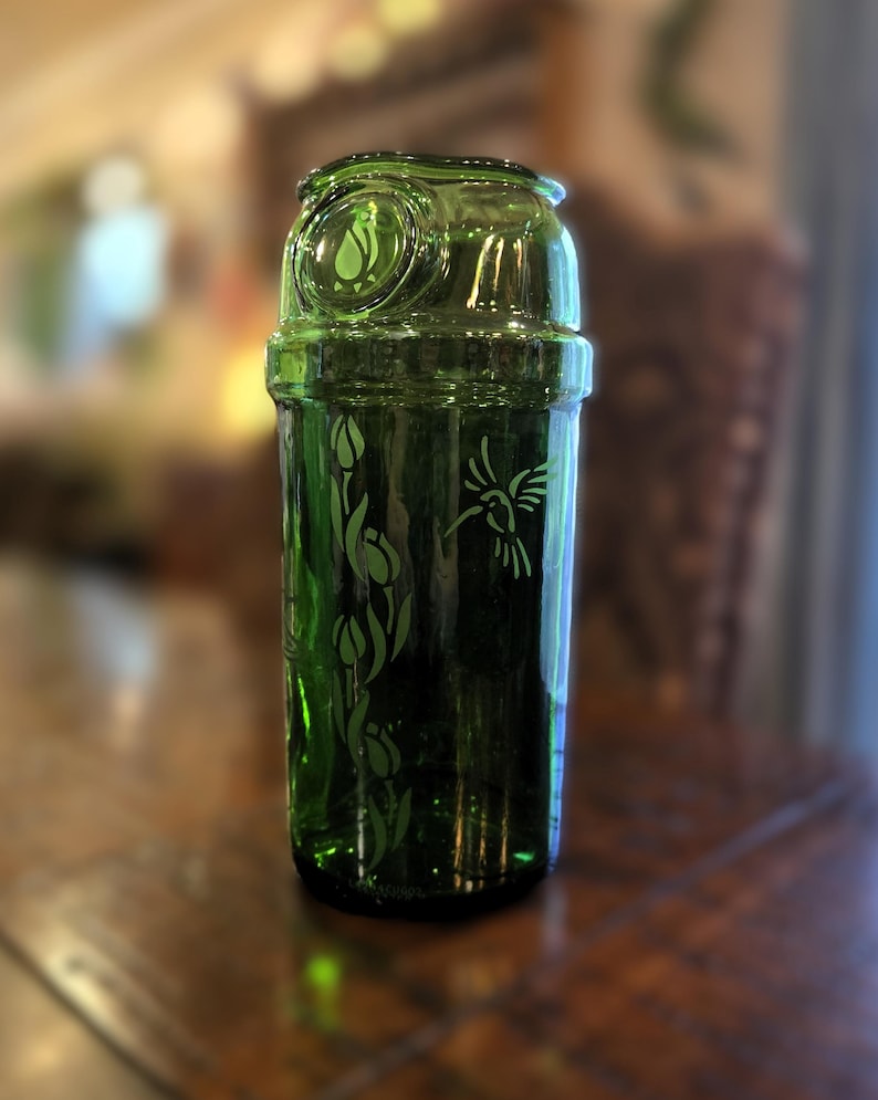 May include: A green glass bottle with a decorative design. The bottle features etched images of flowers and a hummingbird. The top of the bottle has a circular design with a leaf. The bottle is sitting on a wooden surface.