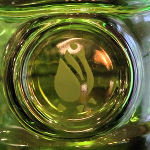 May include: Close-up of a green glass object with a circular design in the center. The design features a stylized flower or leaf shape. The glass has a glossy finish and appears to be a decorative item.