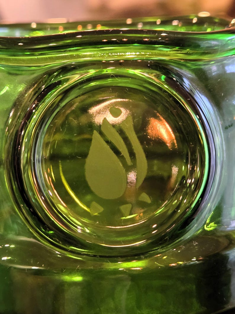 May include: Close-up of a green glass object with a circular design in the center. The design features a stylized flower or leaf shape. The glass has a glossy finish and appears to be a decorative item.