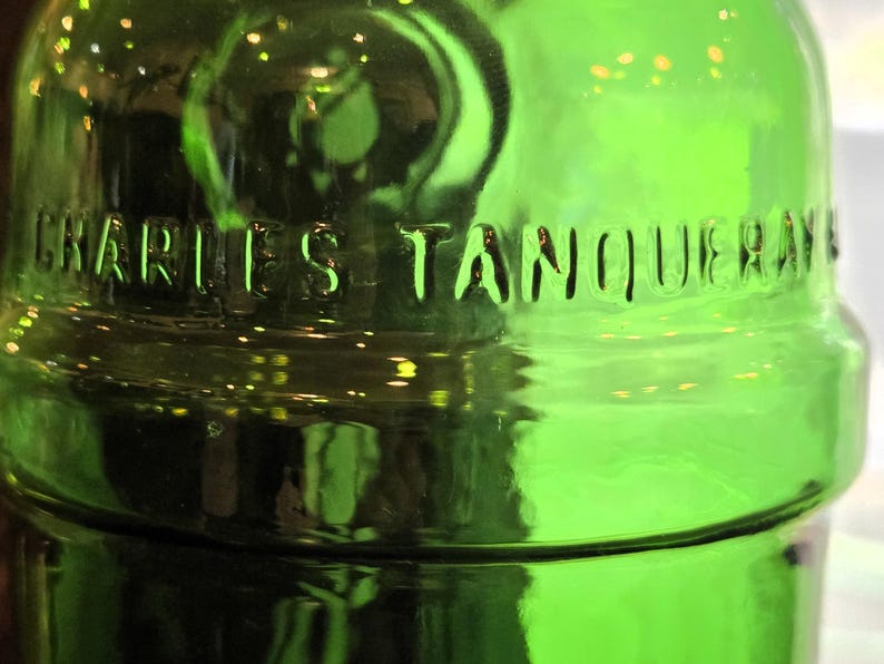 May include: Close-up of a green glass bottle with the embossed text "CHARLES TANQUERAY". The bottle has a rounded shape and a glossy finish, reflecting light. The overall theme is related to alcoholic beverages.
