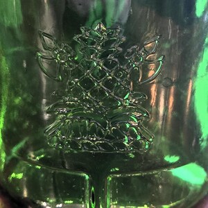 May include: Close-up of a green glass bottle with a raised, detailed pine tree design. The bottle's surface reflects light, highlighting the embossed texture of the tree and the surrounding glass. The design is centered on the bottle.