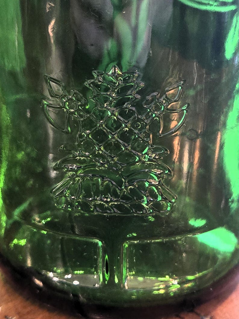 May include: Close-up of a green glass bottle with a raised, detailed pine tree design. The bottle's surface reflects light, highlighting the embossed texture of the tree and the surrounding glass. The design is centered on the bottle.