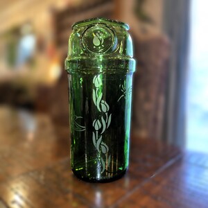 May include: A green glass bottle with a decorative floral design. The bottle has a cylindrical shape with a rounded top and a detailed etched pattern of tulips. The glass has a slight tint, and the design is etched in a lighter color.