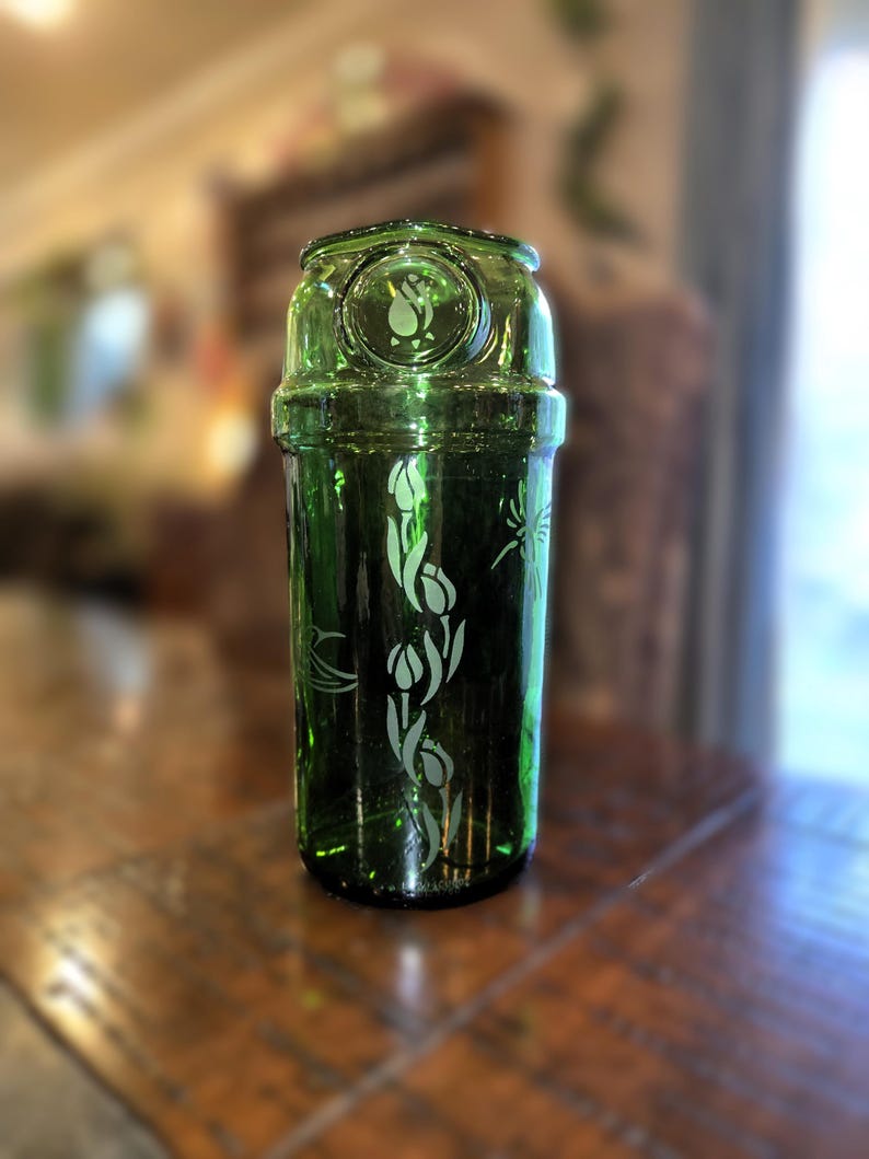 May include: A green glass bottle with a decorative floral design. The bottle has a cylindrical shape with a rounded top and a detailed etched pattern of tulips. The glass has a slight tint, and the design is etched in a lighter color.