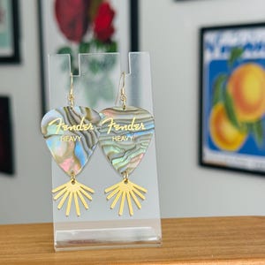 May include: A pair of guitar pick earrings with a pearlescent finish. The earrings have the Fender logo and the word "HEAVY" printed in gold. The earrings are suspended from gold-colored hooks and feature a gold sunburst design.