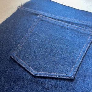 May include: Close-up of a section of dark blue denim fabric. The image highlights a back pocket with orange stitching. The pocket is rectangular with a pointed bottom. The denim has a textured appearance.