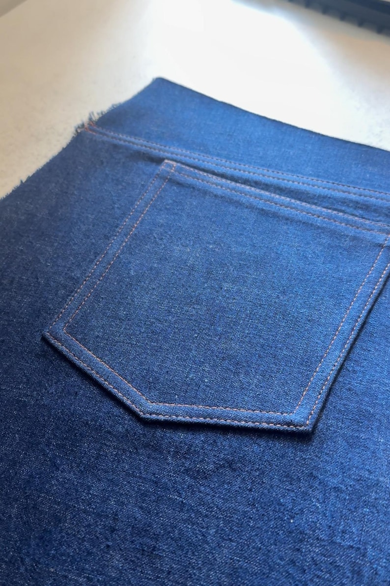 May include: Close-up of a section of dark blue denim fabric. The image highlights a back pocket with orange stitching. The pocket is rectangular with a pointed bottom. The denim has a textured appearance.