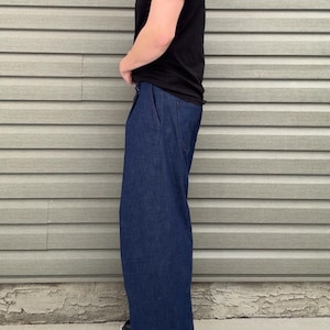 May include: A person wearing a black t-shirt and dark blue denim pants. The pants have a wide leg design. The person is wearing black boots. The background is a light gray siding.
