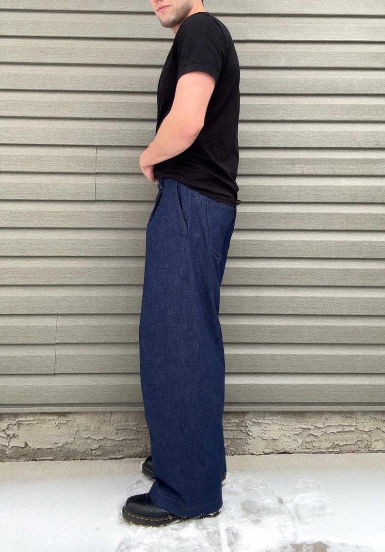 May include: A person wearing a black t-shirt and dark blue denim pants. The pants have a wide leg design. The person is wearing black boots. The background is a light gray siding.