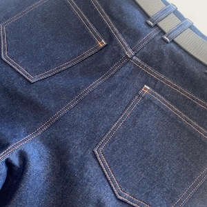 May include: Close-up of the back of dark blue denim jeans with orange stitching. The jeans have two back pockets and a gray belt. The stitching and belt loops are visible.