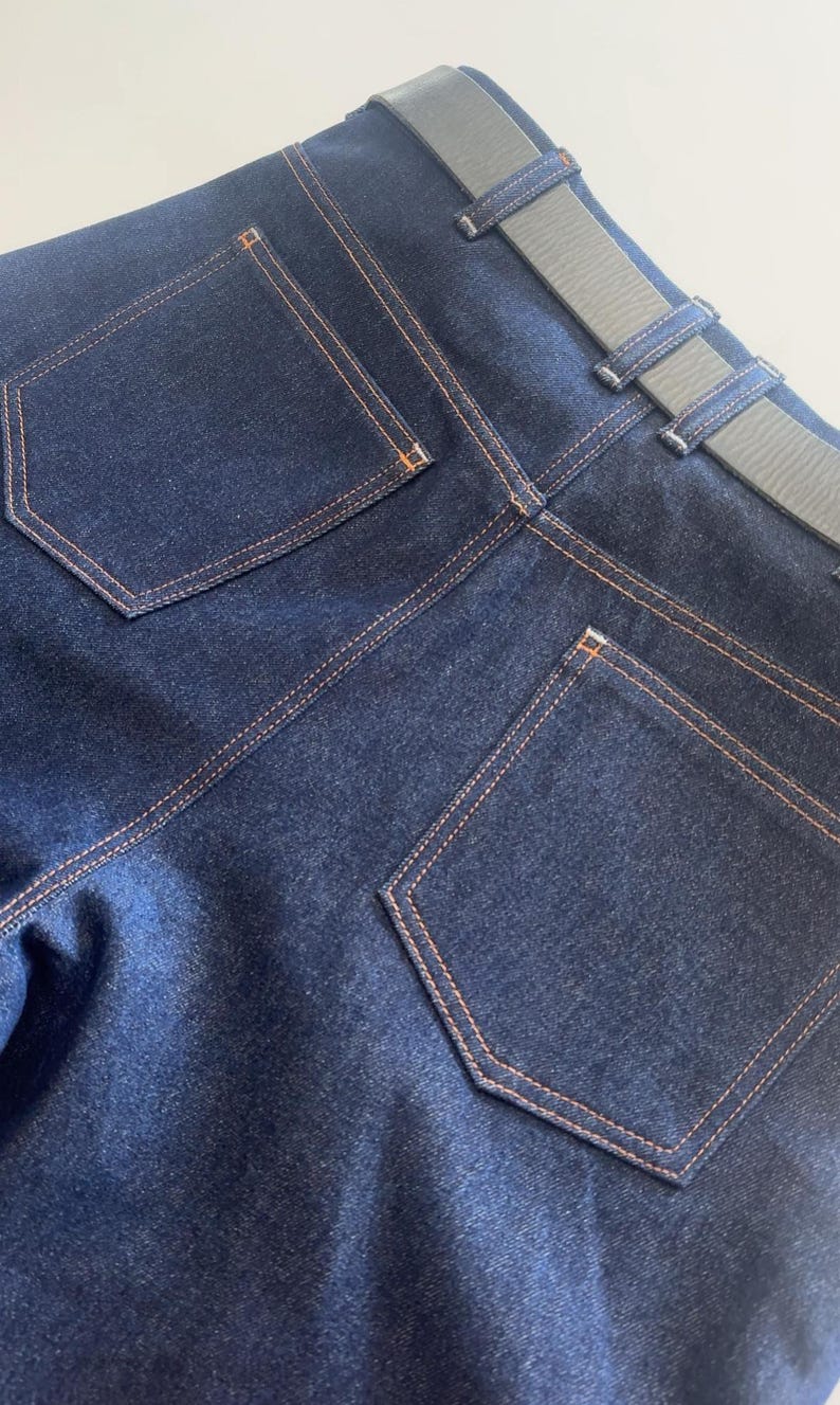 May include: Close-up of the back of dark blue denim jeans with orange stitching. The jeans have two back pockets and a gray belt. The stitching and belt loops are visible.
