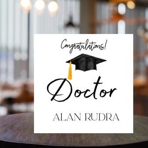 Puede incluir: Una tarjeta de graduación blanca con las palabras "Congratulations!" y "Doctor" en escritura negra. Un birrete negro con una borla dorada está encima de la palabra "Doctor". El nombre "ALAN RUDRA" está impreso debajo.