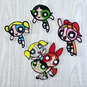 May include: A collection of five embroidered patches featuring cartoon characters with large eyes. The patches depict figures with black, blonde, and red hair, wearing green, blue, and pink outfits, rendered in vibrant thread colors on a white background.